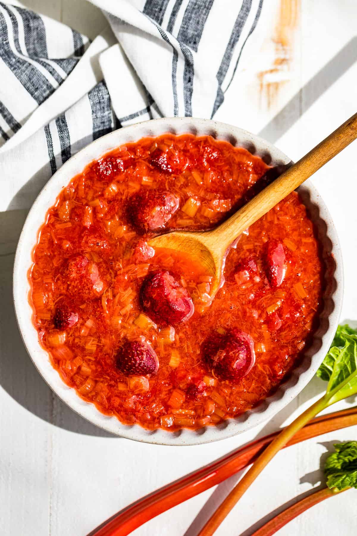 Strawberry Rhubarb Sauce in a grey pottery bowl with a wooden spoon and a blue and white linen on the side with 3 stalks of rhubarb on the other side.