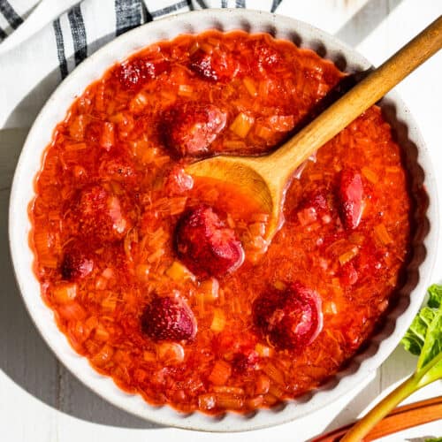 Strawberry Rhubarb Sauce in a grey pottery bowl with a wooden spoon and a blue and white linen on the side with 3 stalks of rhubarb on the other side.