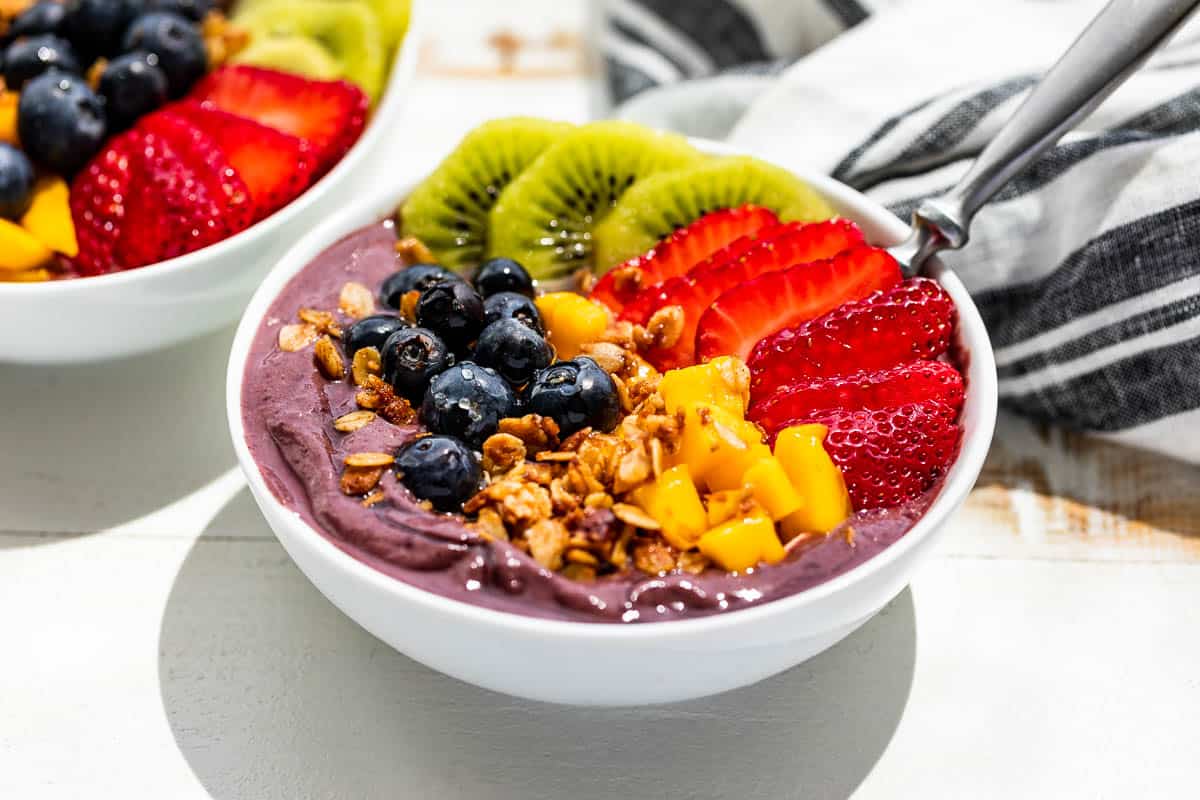 Side view of two acai bowls topped with granola and fresh fruit on a white background with a blue and white linen in the background.