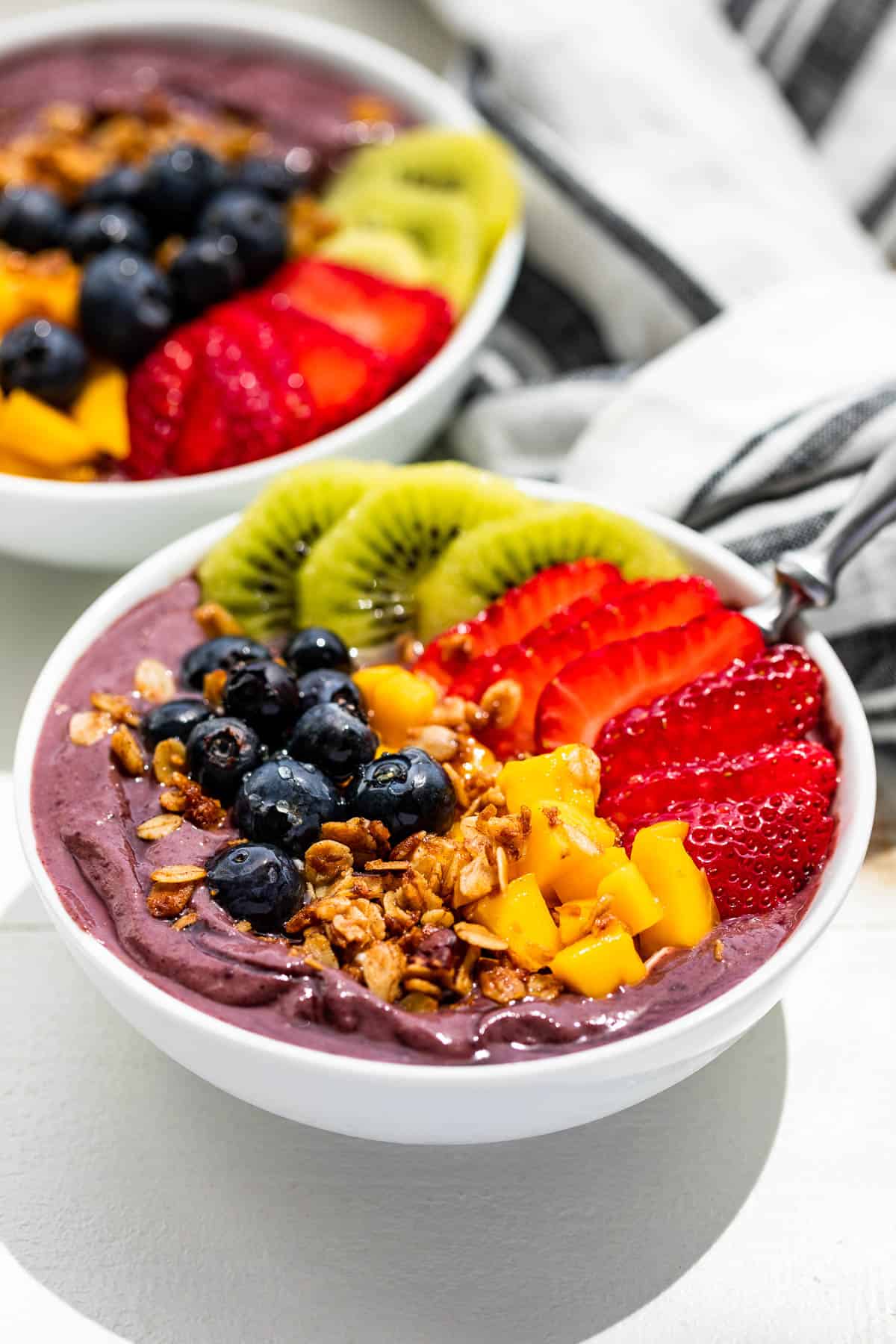 Side view of two white bowls with an acai smoothie base topped with granola, berries, and diced mango.