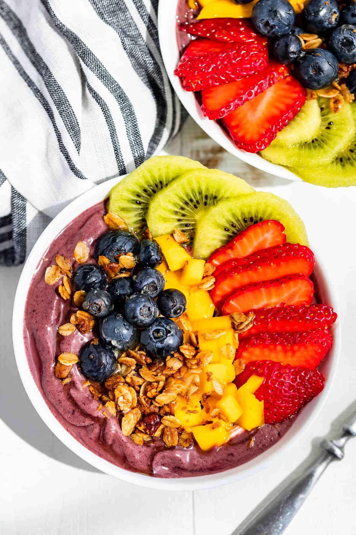 Straight down view of two white bowls filled with acai smoothie base topped with strawberries, sliced kiwi, blueberries and granola.
