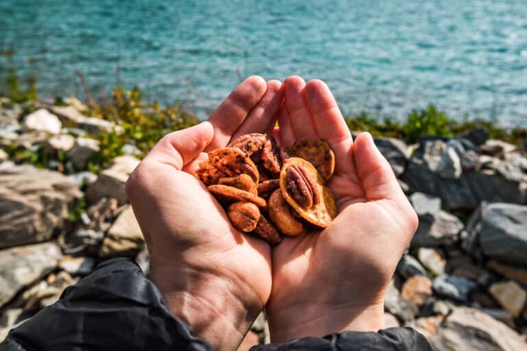 Hands holding Honey Mustard Trail Mix with rocks and an aqua blue lake in the background.