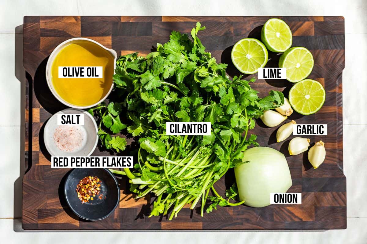 All the ingredients for Cilantro Chimichurri on a wood cutting board: cilantro, olive oil, garlic, onion, lime, red peppers flakes, and sea salt.