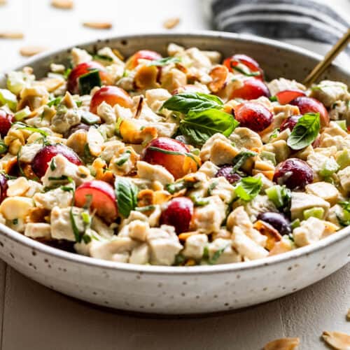 Chicken salad in a speckled pottery bowl on a white background with sliced almonds around it and a blue and white linen behind the bowl.