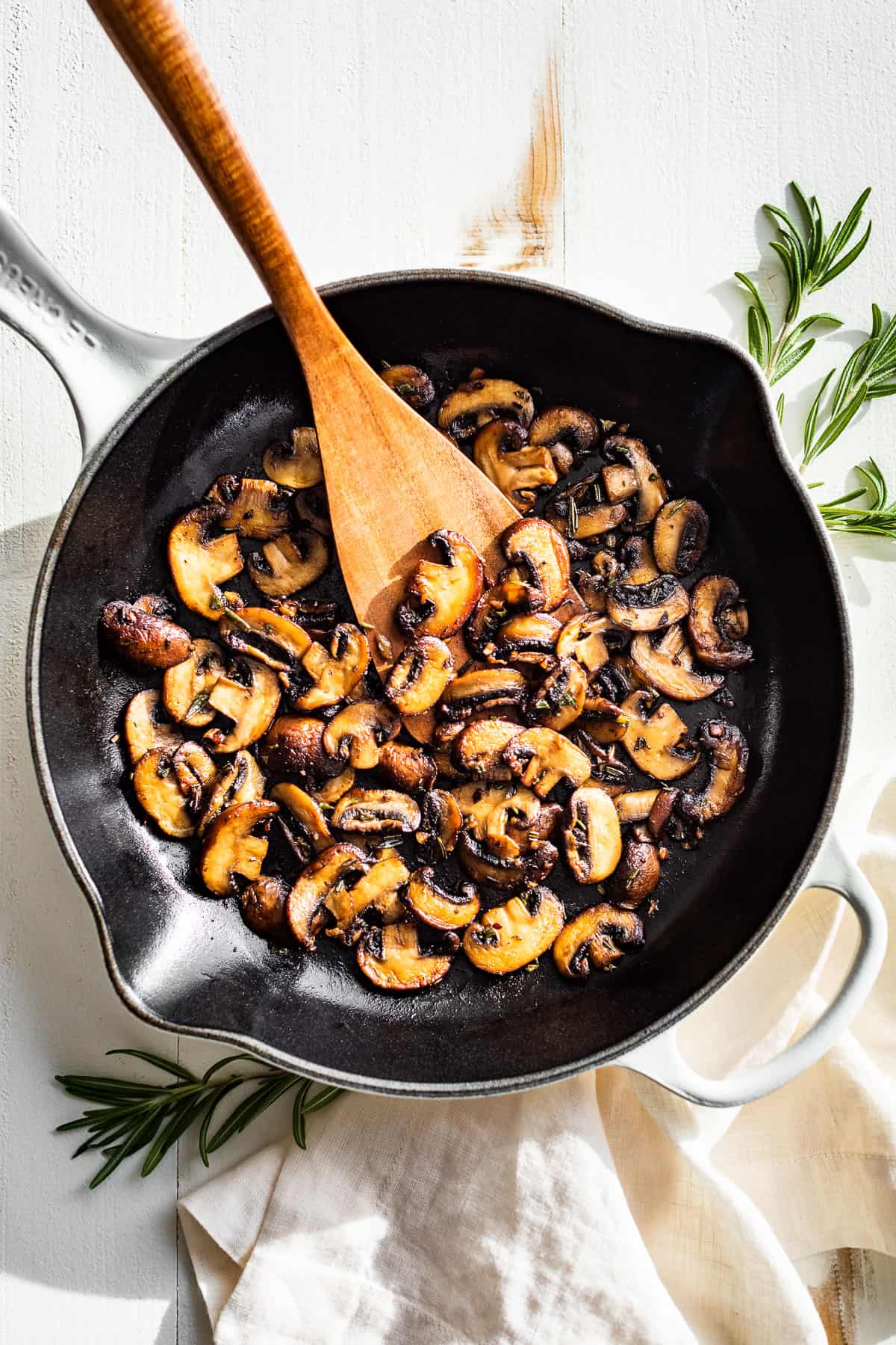 Straight down view of garlic butter mushrooms in a white skillet with a wood spatula and rosemary sprigs on the side.
