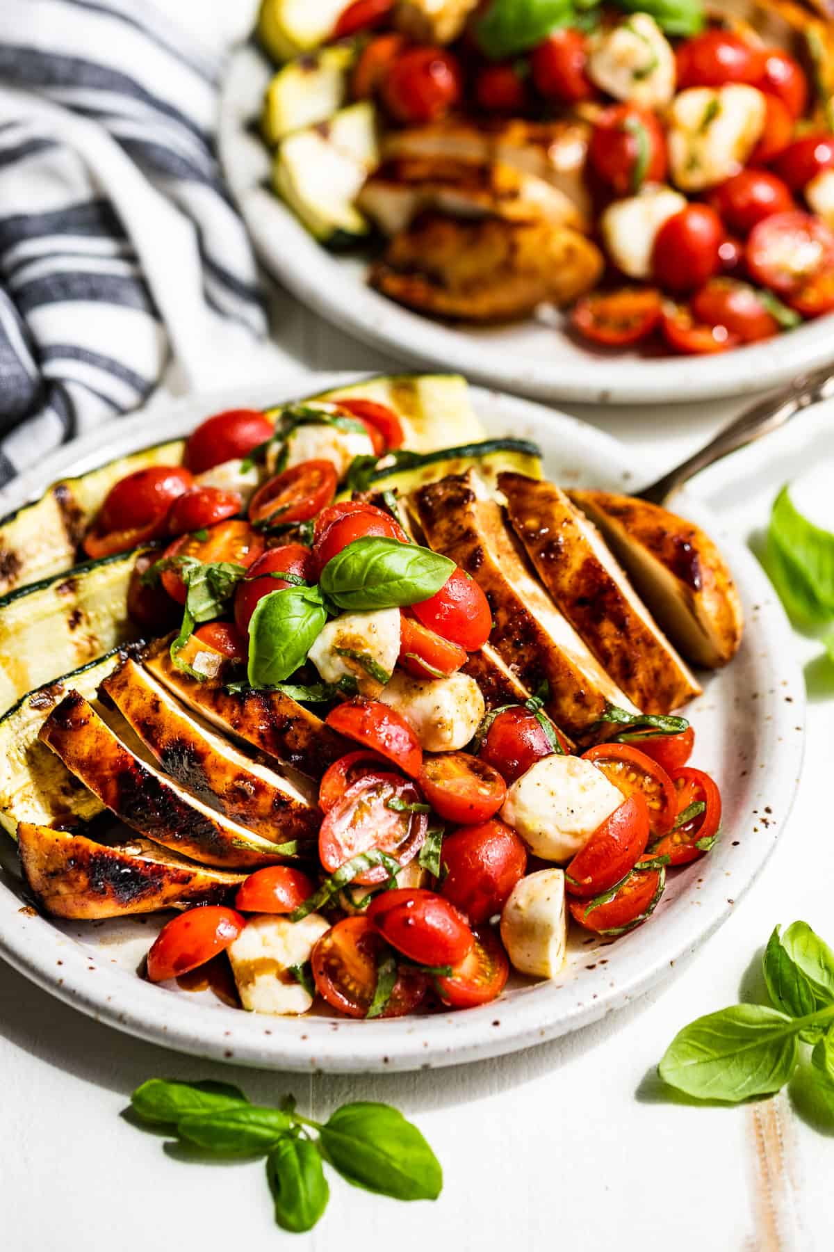 Sliced balsamic chicken topped with caprese salad and zucchini on the side on a pottery plate.