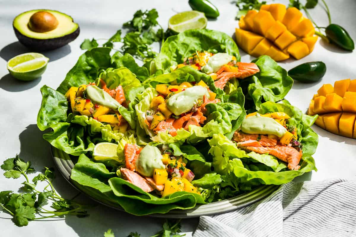 Fish taco lettuce wraps on a round silver platter with mangos and avocados in the background.