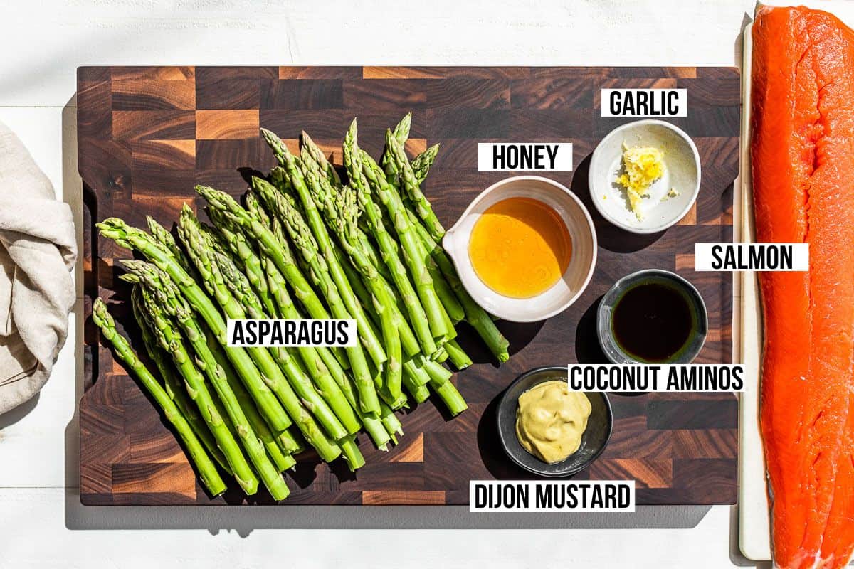 Asparagus, salmon filet, honey, Dijon mustard, garlic, and coconut aminos in containers on a wood cutting board.