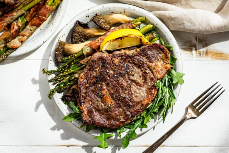 Straight down view of a grilled ribeye steak on a bed of arugula with asparagus and mushrooms on the side.