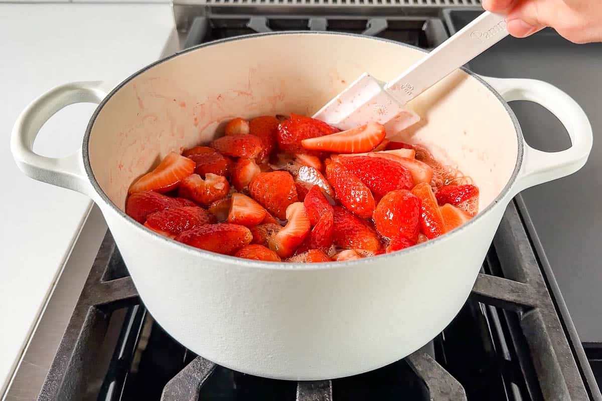 Stirring the strawberries in a large white pot with a spatula as it comes to a boil.