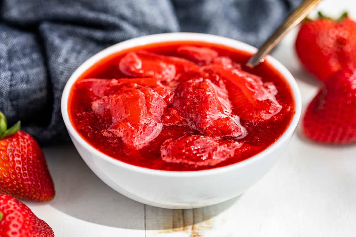 Side view of Strawberry Compote in a white bowl with strawberries on the side and blue linen in the background.