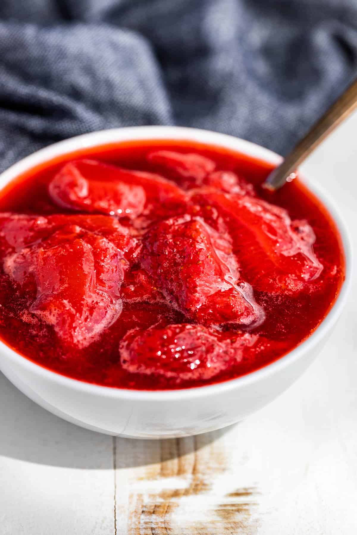 Side view of Strawberry Compote in a white bowl with a silver spoon in it and a blue linen in the background.