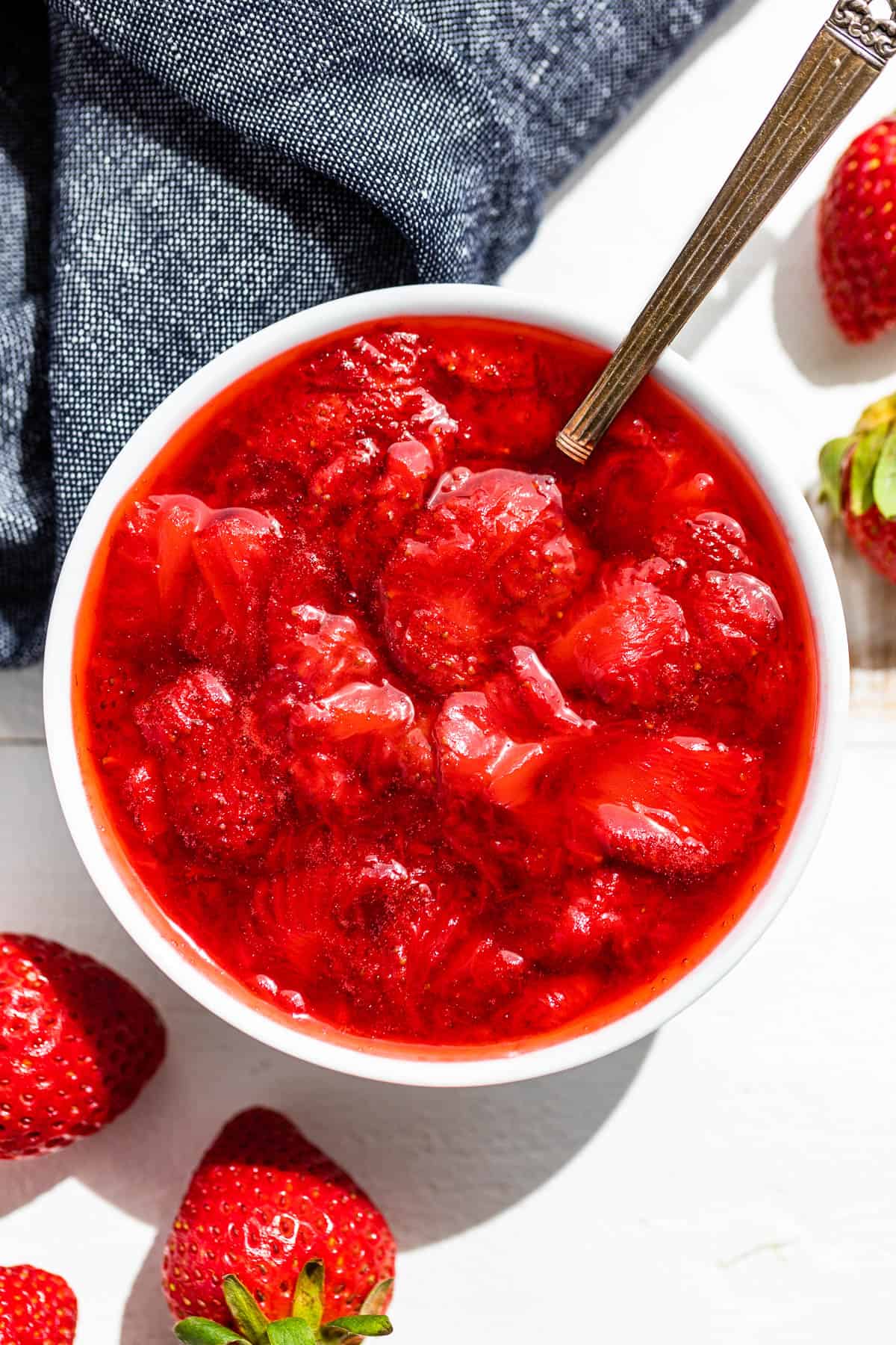 Straight down view of Strawberry Compote in a white bowl with a silver spoon and strawberries on the side with a blue linen in the background.