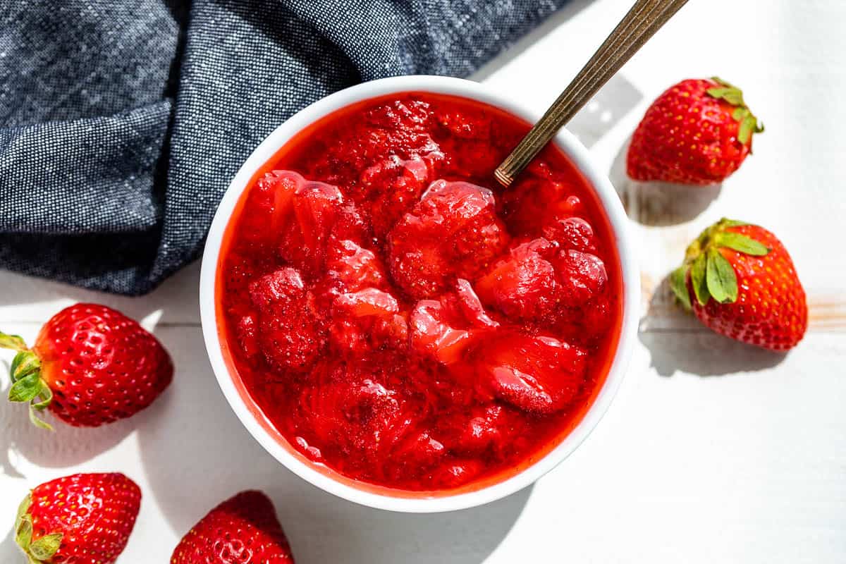 Straight down view of Strawberry Compote in a white bowl with strawberries and a blue linen on the side.