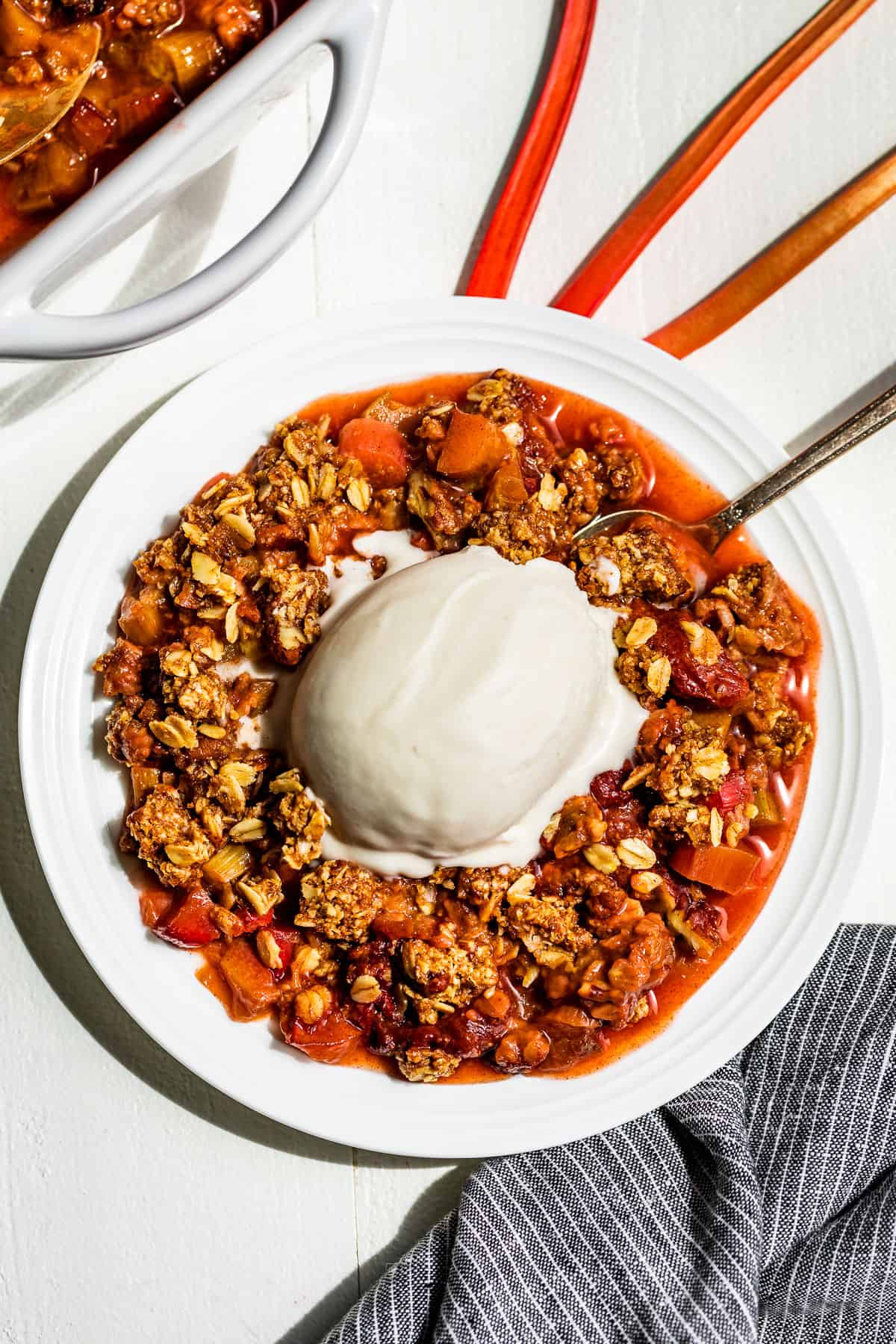 A white plate with Strawberry Rhubarb Crisp with a scoop of ice cream on top with a blue linen on the side.
