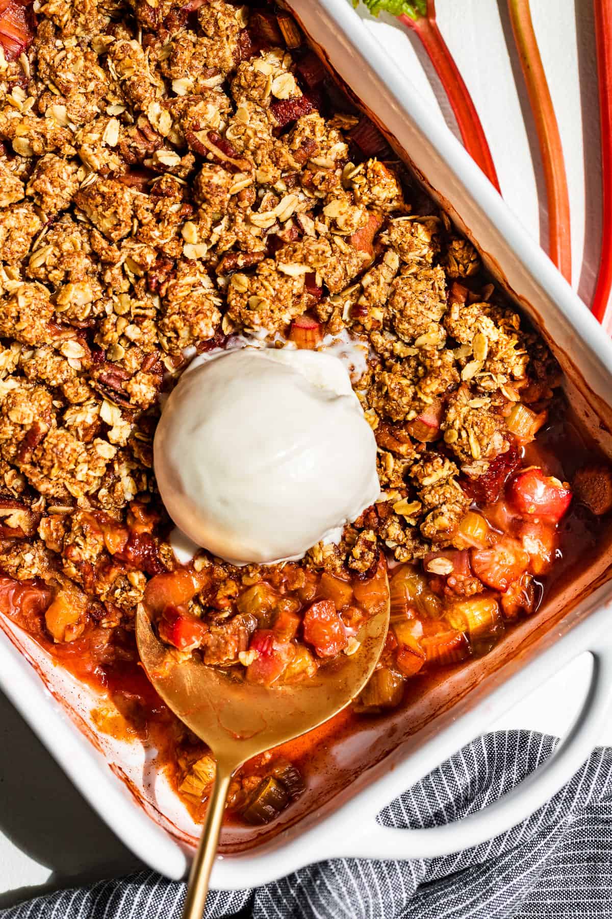 Strawberry Rhubarb Crisp in a white baking dish with a large scoop of ice cream and a gold serving spoon.