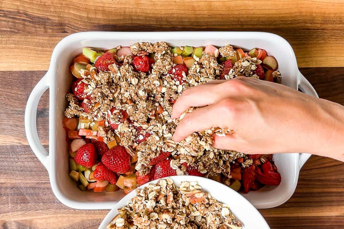 Adding the crisp topping over the top of the rhubarb and strawberries in a white baking dish.