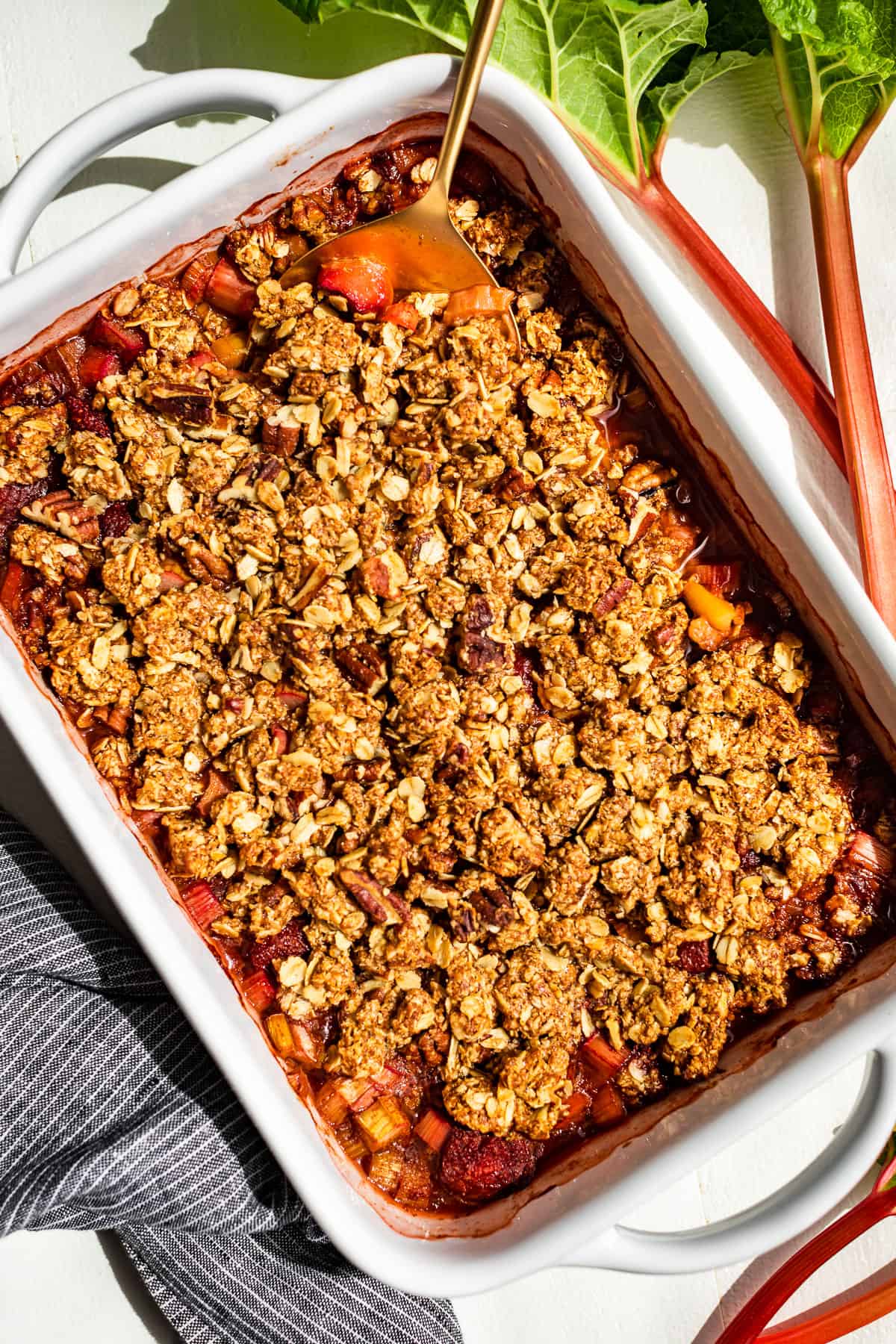 Straight down view of strawberry rhubarb crisp in a white baking dish on an angle with stalks of rhubarb on the side.