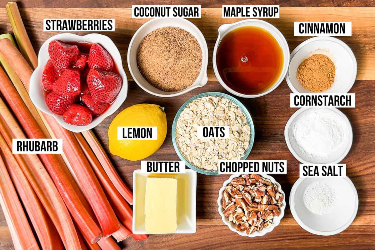 All the ingredients for Strawberry Rhubarb Crisp: rhubarb stalks, strawberries, maple syrup, coconut sugar, butter, oats, chopped nuts, cinnamon, lemon, cornstarch, and sea salt in small bowls on a wood cutting board.