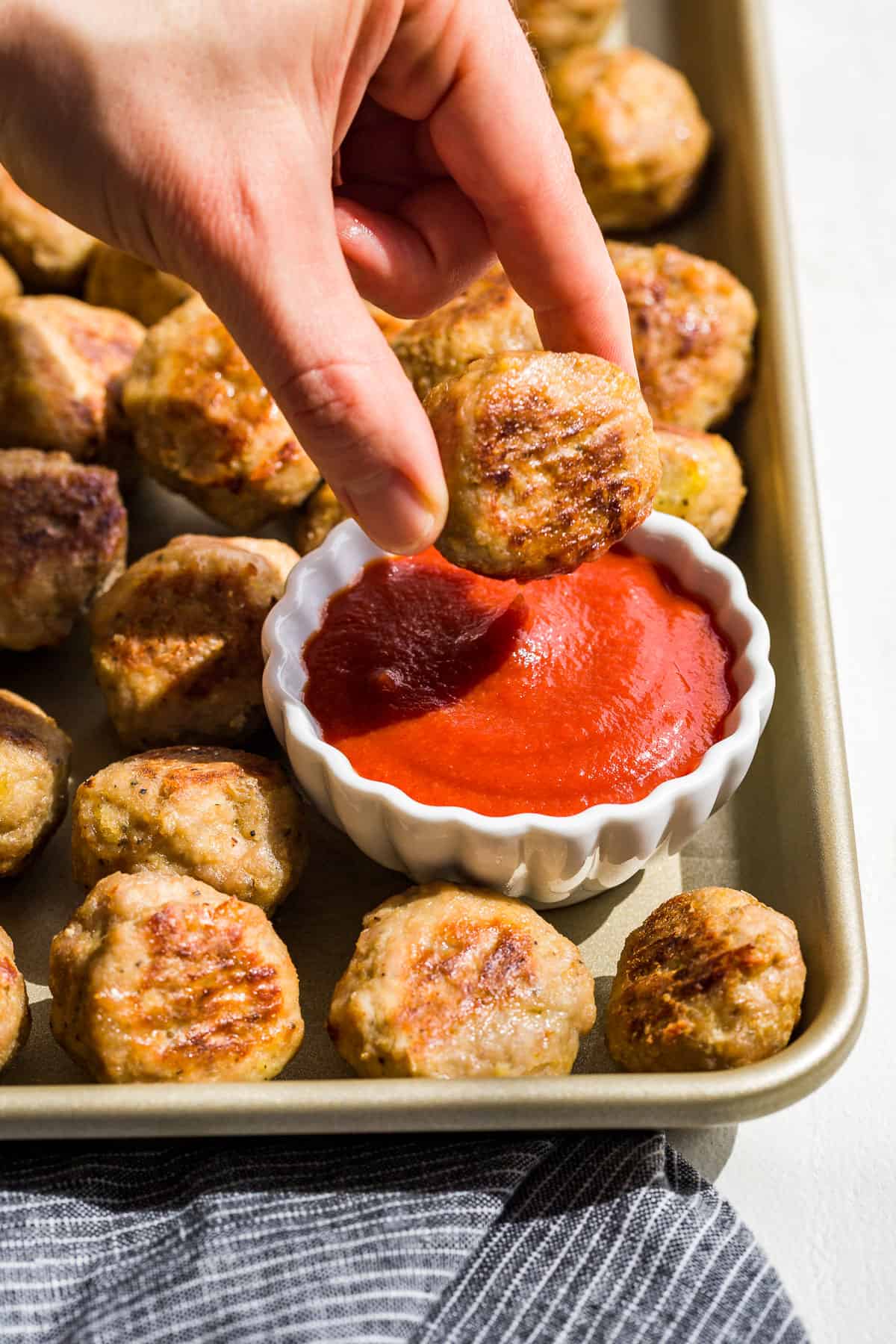 Side view of Baked Turkey Meatballs on a gold baking sheet with a hand dunking one meatball into a small bowl of ketchup.