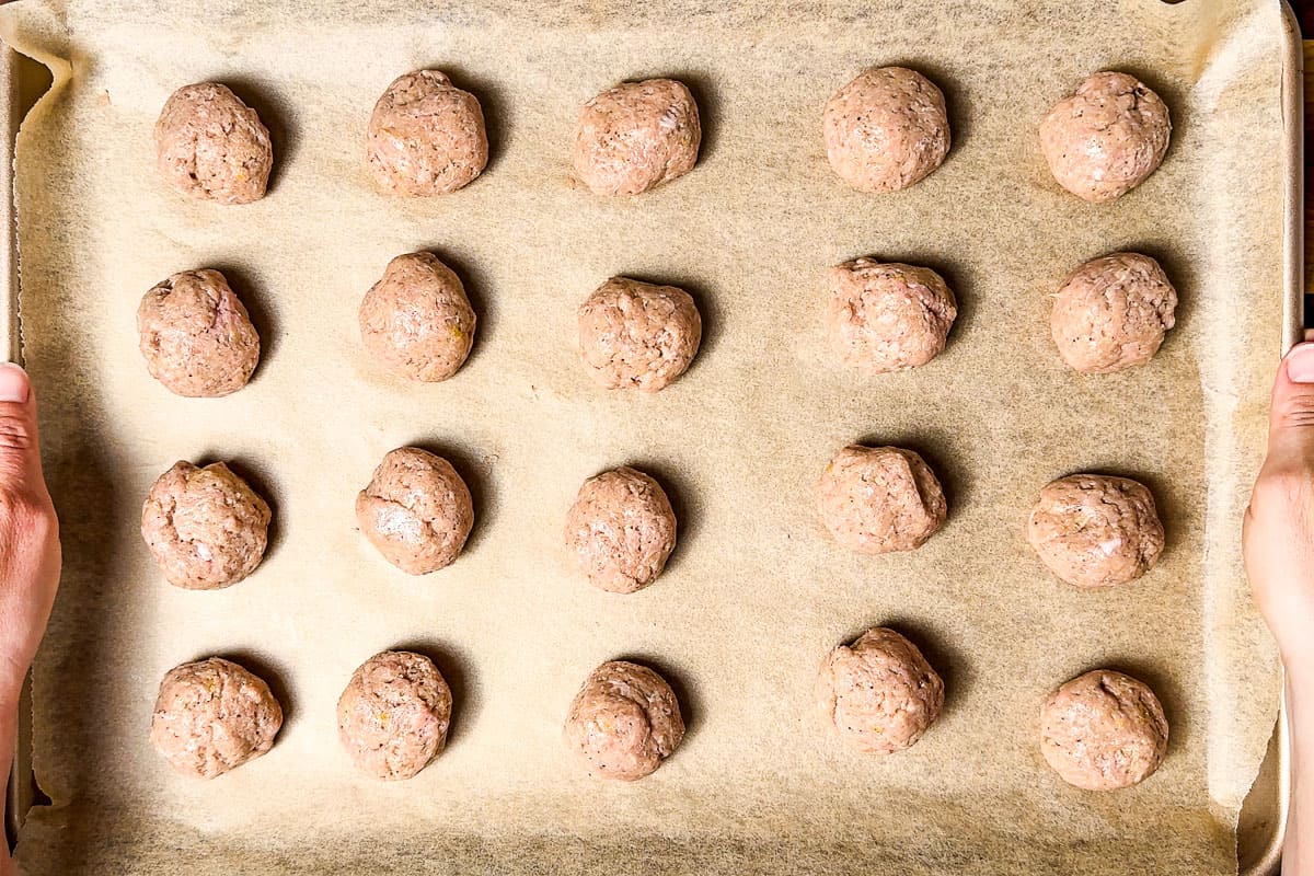 Rolled turkey meatballs on a parchment lined baking sheet with two hands lifting it up.