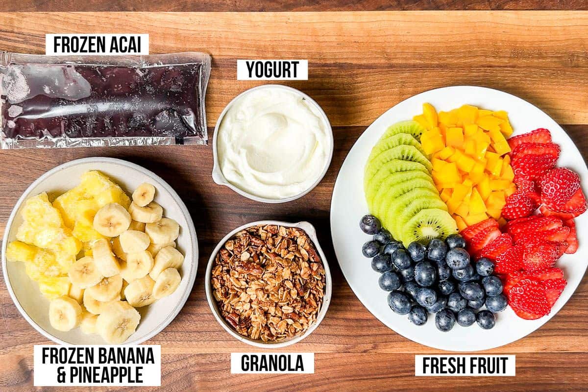 Frozen acai puree, frozen bananas and pineapple, granola, yogurt, fresh kiwi, blueberries, strawberry, and mango on a wood cutting board.