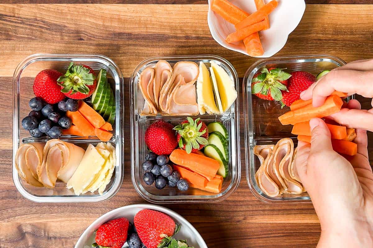 Adding carrot sticks to the Adult Lunchables containers on a wood cutting board.