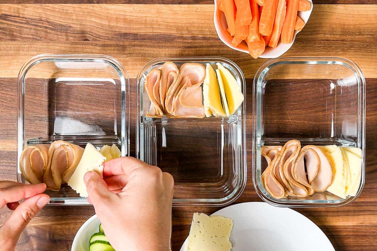 Placing sliced turkey and sliced cheese into glass meal prep containers on a wood cutting board.