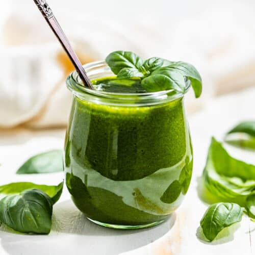 Side view of finished Basil Vinaigrette in a glass tulip shaped jar with basil leaves around it.