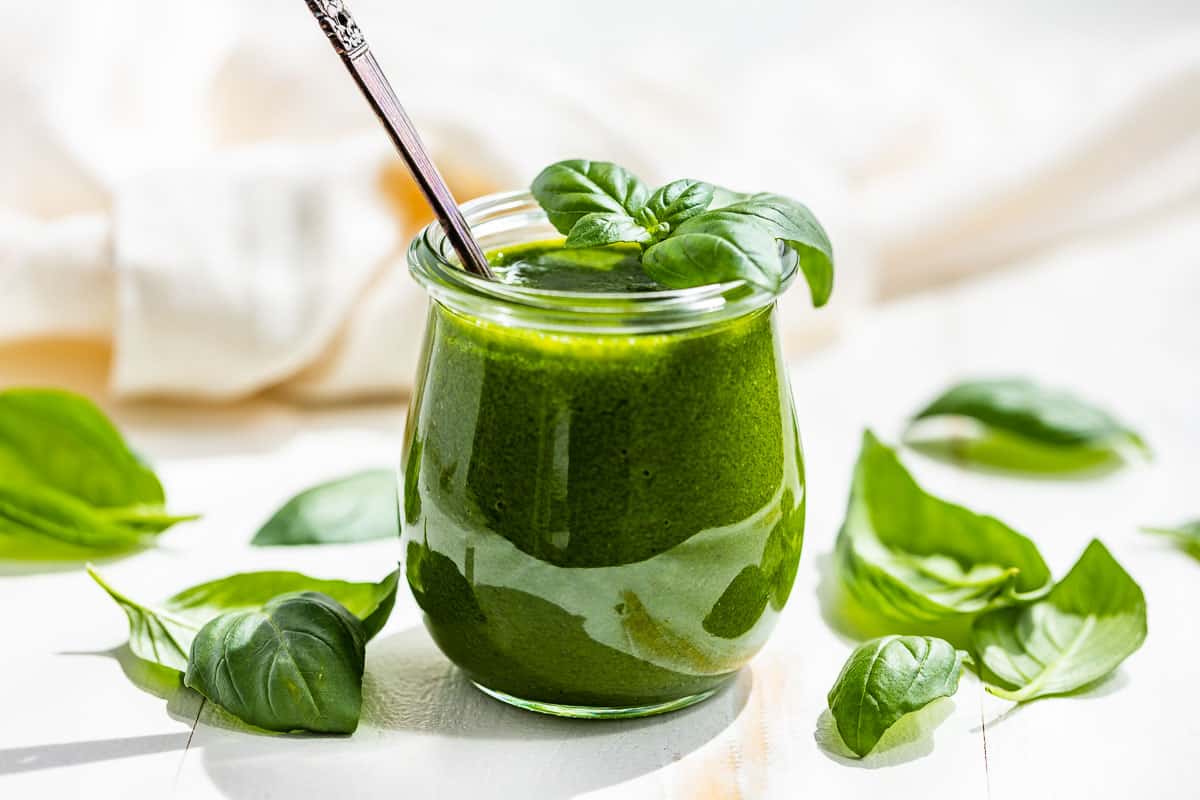 Side view of finished Basil Vinaigrette in a glass tulip shaped jar with basil leaves around it.