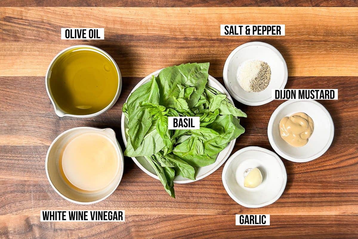 Olive oil, white wine vinegar, fresh basil, garlic, Dijon mustard, salt and pepper in small pottery bowls on a wood cutting board.
