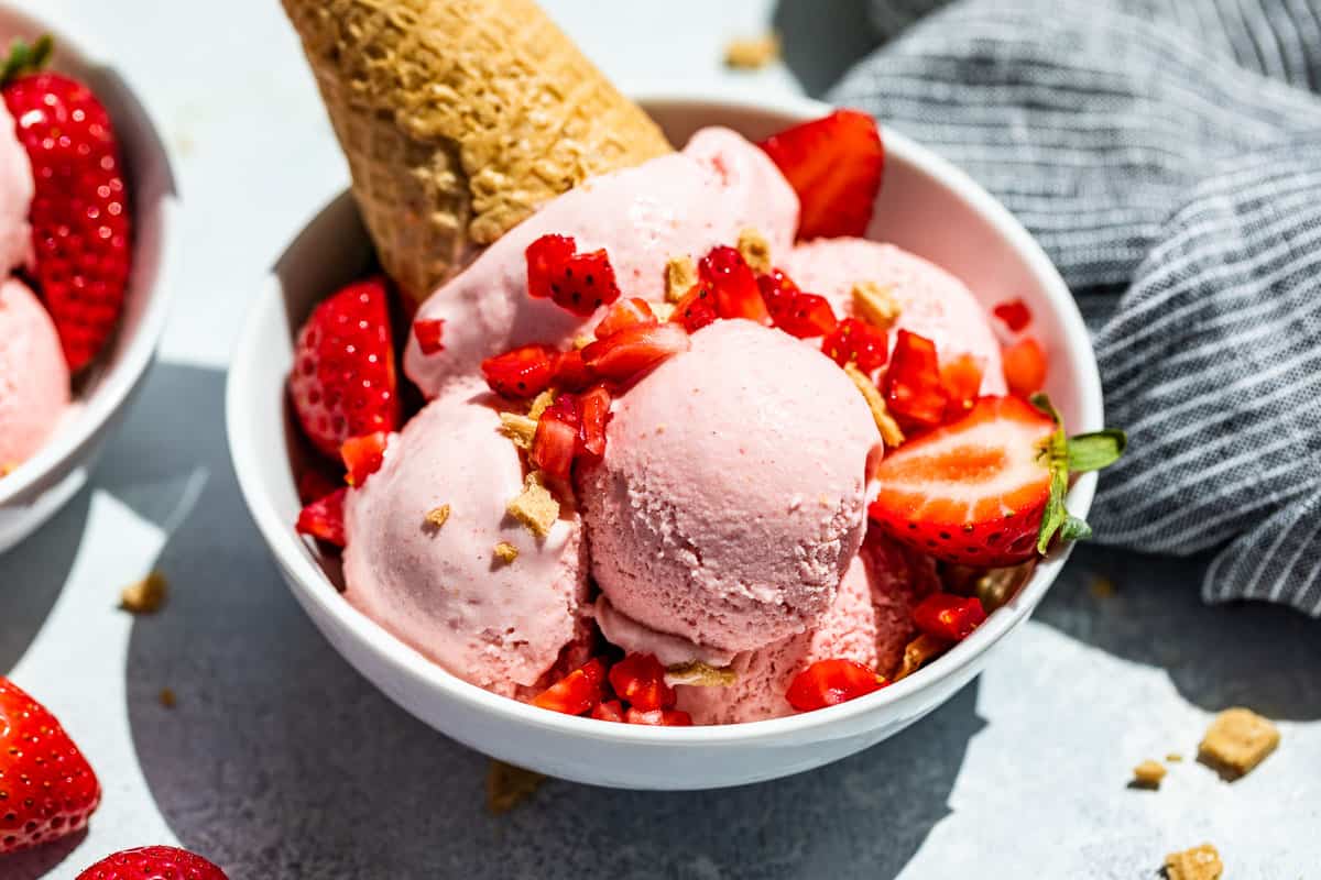 Side view of Strawberry Cheesecake Ice Cream in a white bowl topped with fresh strawberries and a waffle cone on the side.