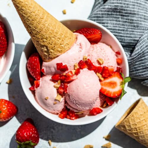 Straight down view of Strawberry Cheesecake Ice Cream scooped into two white bowls topped with strawberries and waffle cones.