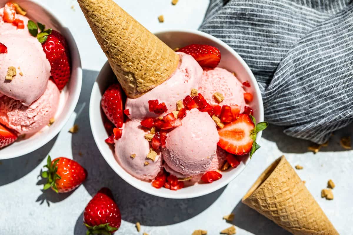 Straight down view of Strawberry Cheesecake Ice Cream scooped into two white bowls topped with strawberries and waffle cones.