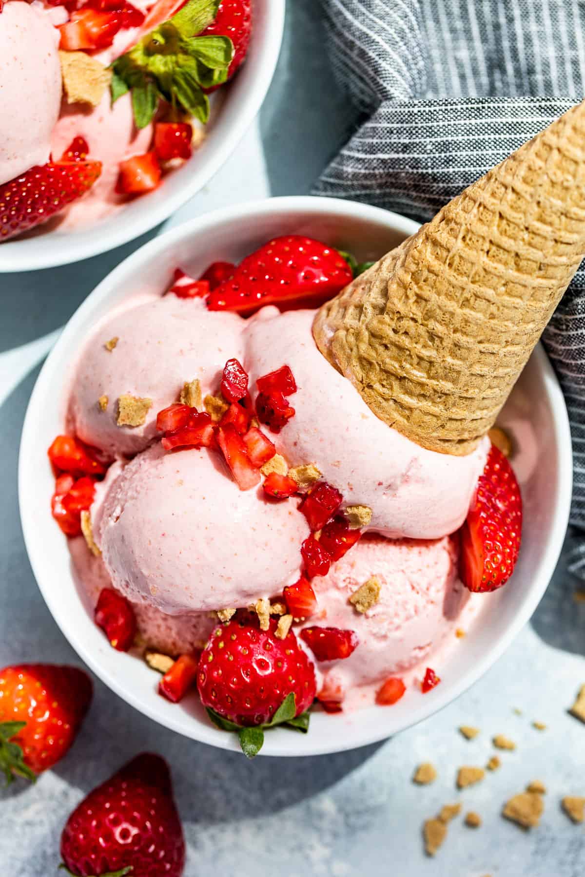 Straight down view of Strawberry Cheesecake Ice Cream scooped into a with bowl and topped with strawberries and a waffle cone on the side.