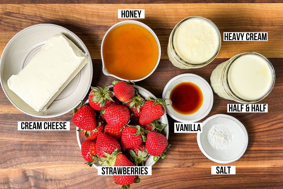 Cream cheese, fresh strawberries, honey, heavy cream, half and half, vanilla, and salt in small pottery bowls on a wood cutting board.