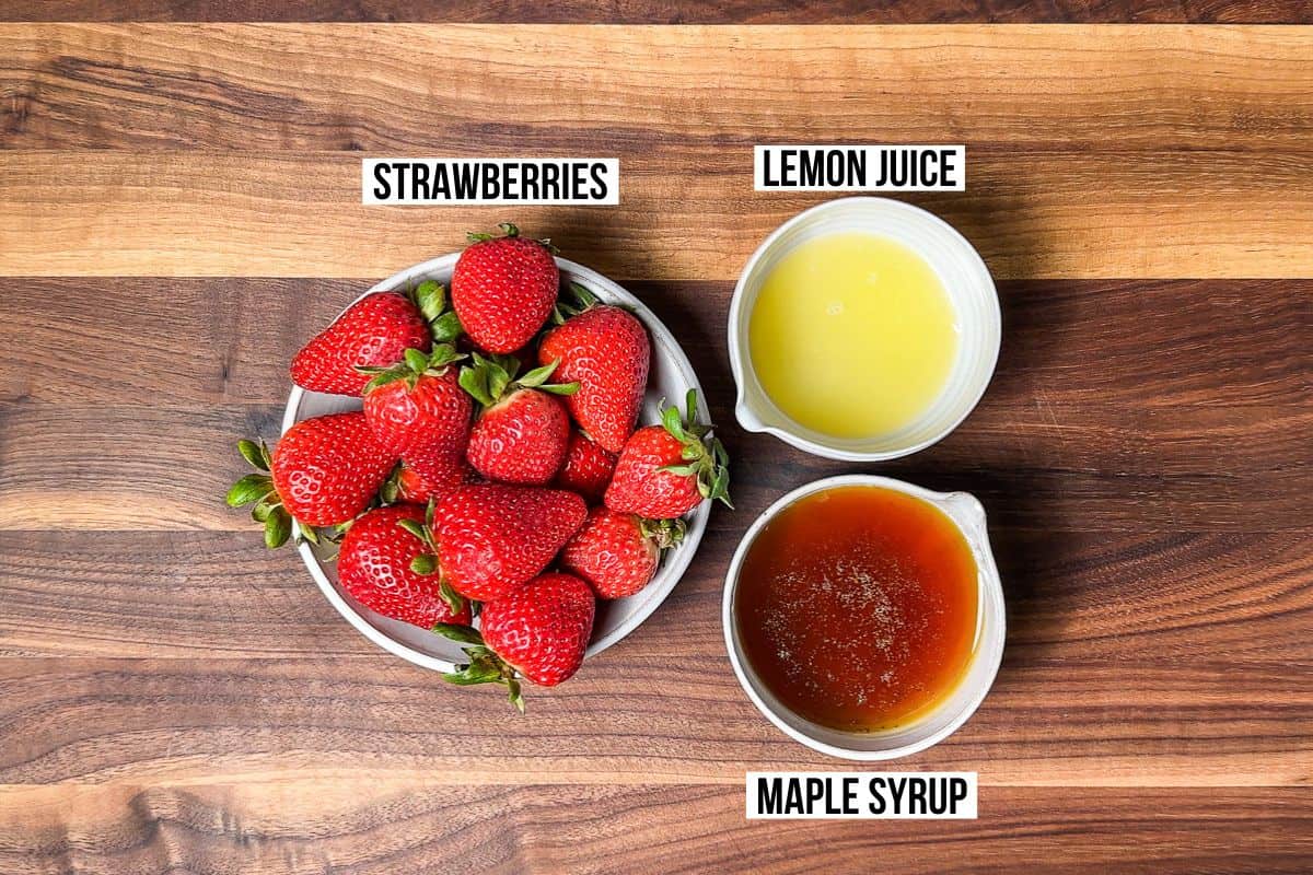 Fresh strawberries, lemon juice, and maple syrup in small pottery bowls on a wood cutting board.