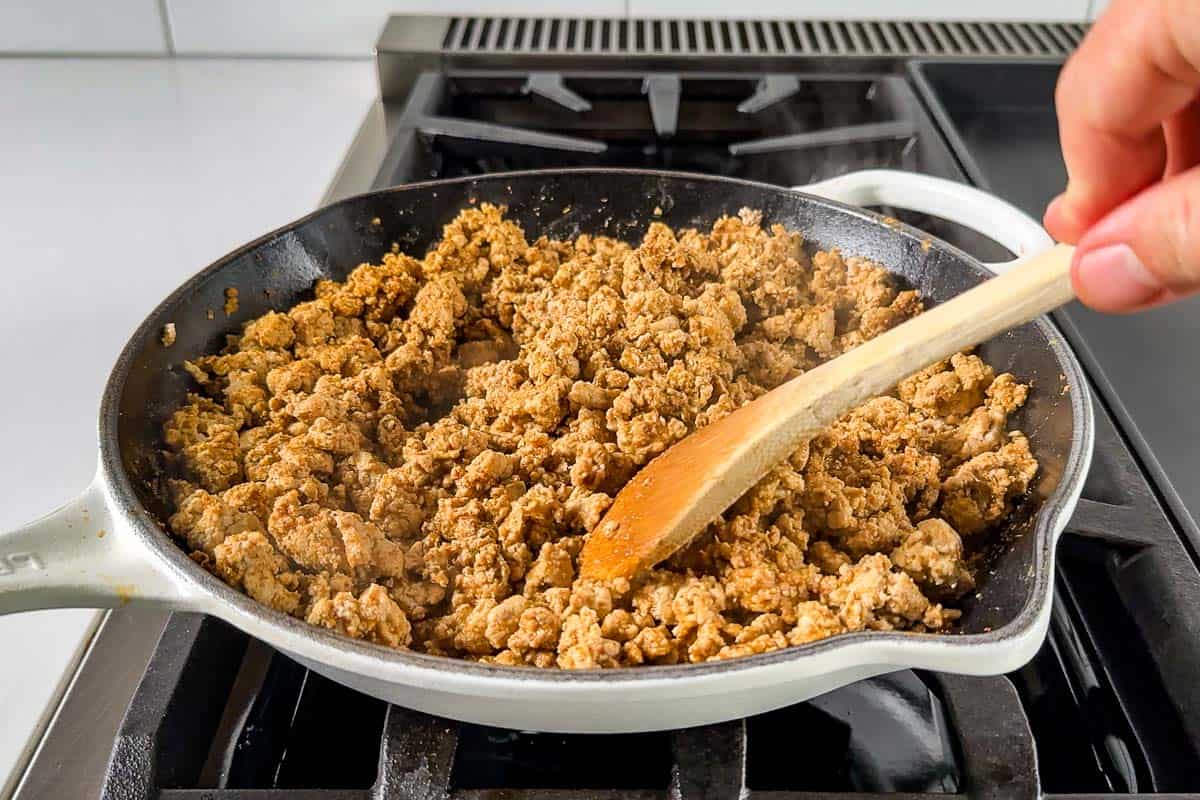 Cooking the ground turkey with taco seasoning in a white skillet with a wood spoon on a stove top.