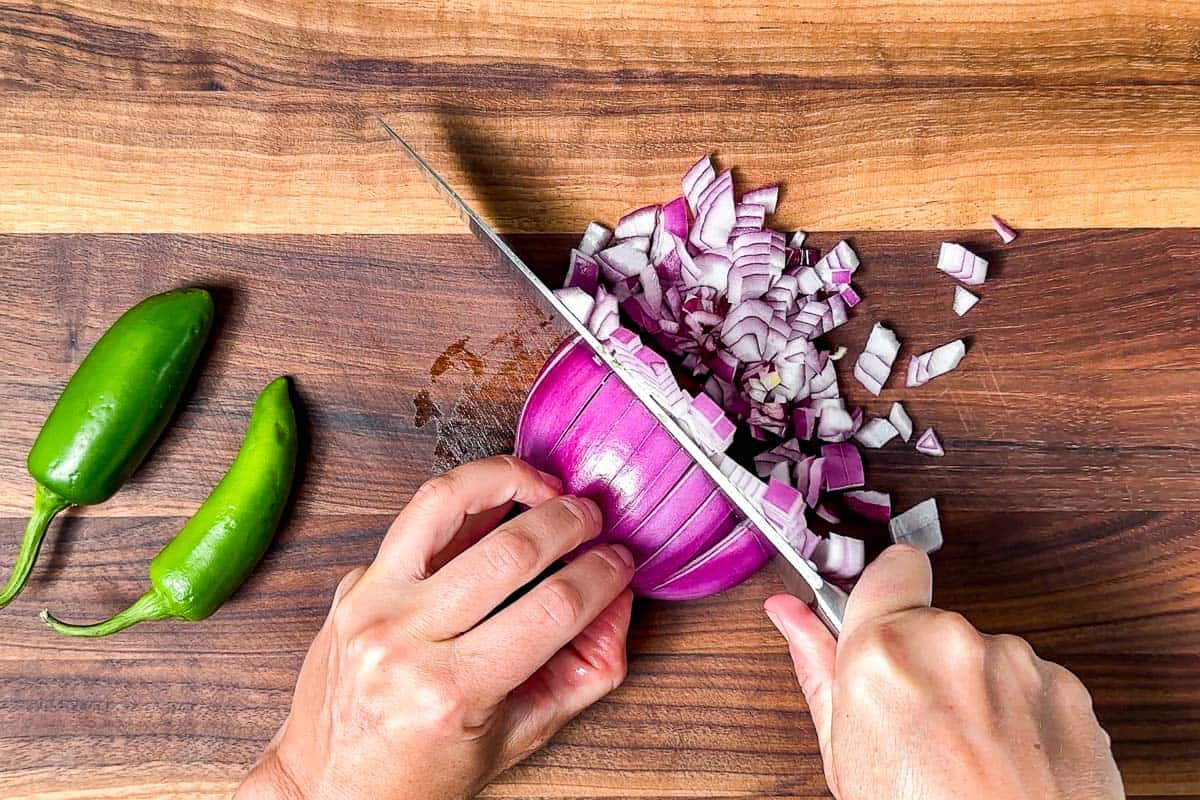 Dicing up red onion on a wood cutting board with jalapenos on the side.