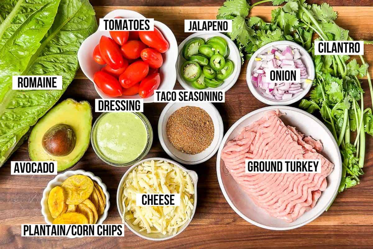 Ground turkey, taco seasoning, avocado, cheese, romaine, cherry tomatoes, onion, cilantro, chips, and fresh cilantro on a wood cutting board.