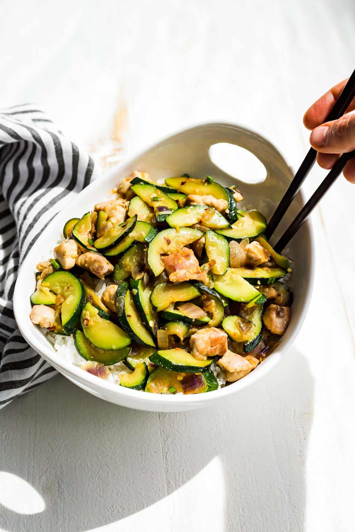 Side view of chicken zucchini stir fry over rice in a white bowl with a hand holding chopsticks scooping some out.