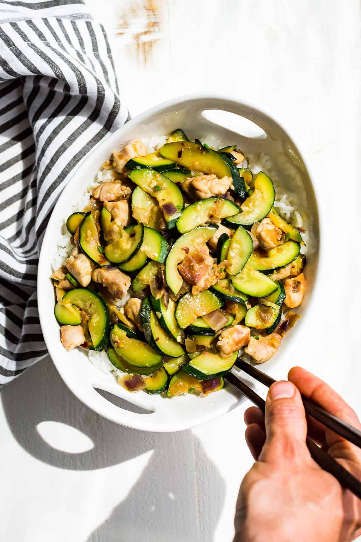 Straight down view of Chicken Zucchini Stir Fry in a bowl over rice with a hand scooping some out with chopsticks.