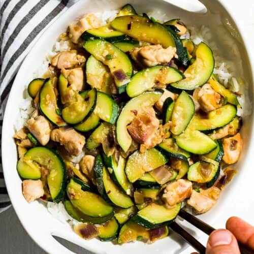 Straight down view of Chicken Zucchini Stir Fry in a bowl over rice with a hand scooping some out with chopsticks.