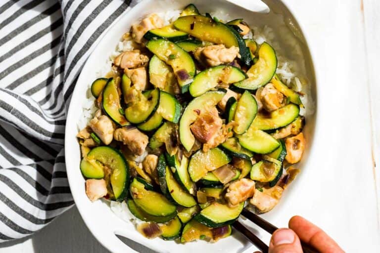 Straight down view of Chicken Zucchini Stir Fry in a bowl over rice with a hand scooping some out with chopsticks.