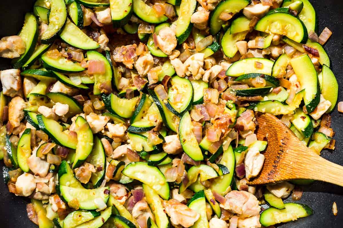 Close up view of the finished Chicken Zucchini Stir Fry in a skillet with a wood spoon in it.