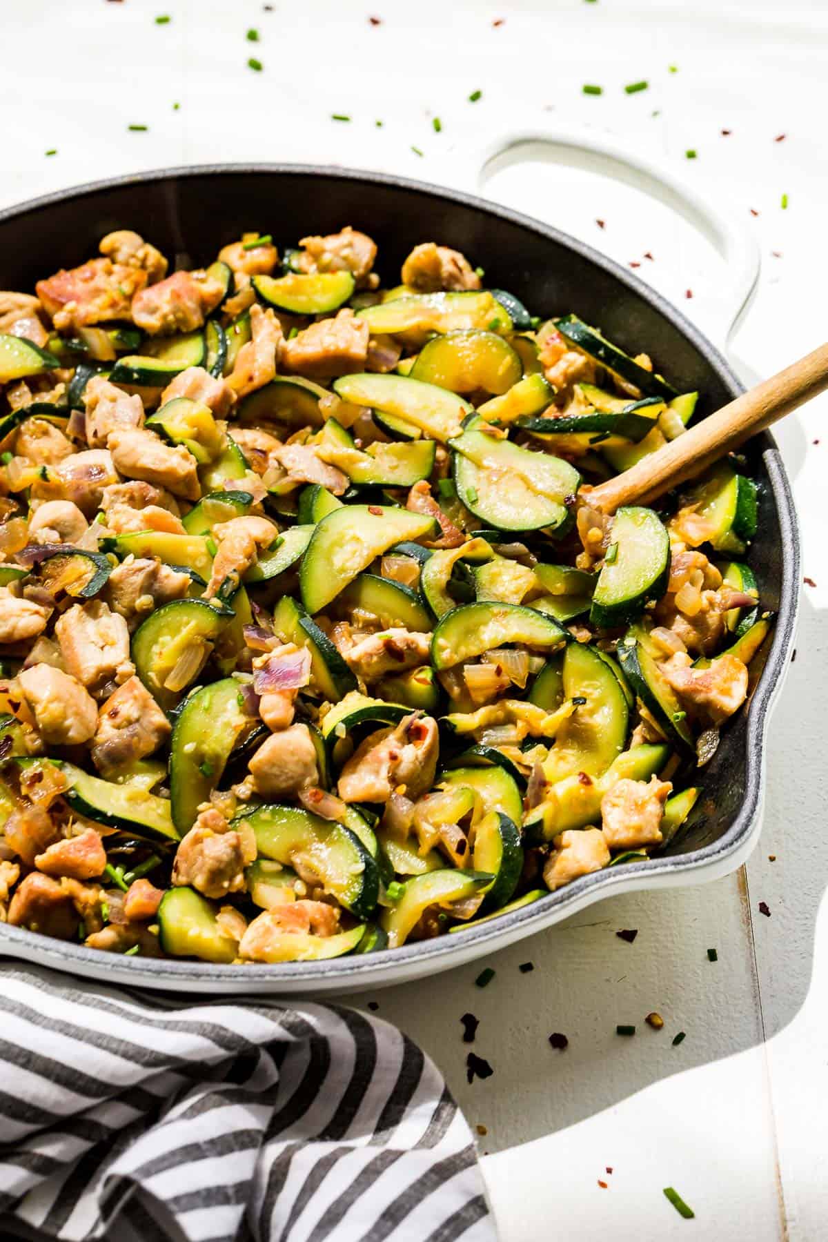 Side view of chicken zucchini stir fry in a white cast iron skillet with a wood spoon in it.