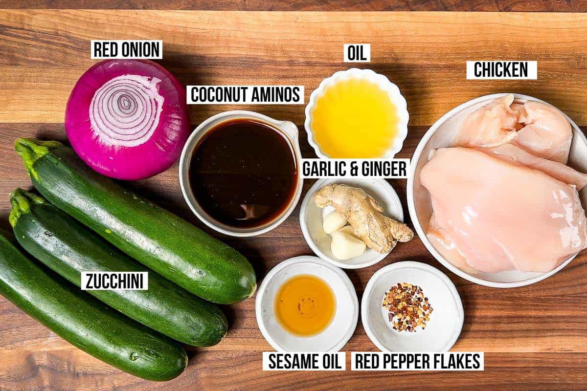 All the ingredients for Chicken Zucchini Stir Fry: zucchini, red onion, chicken, ginger, garlic, sesame oil, coconut aminos, red pepper flakes, and avocado oil in bowls on a wood cutting board.