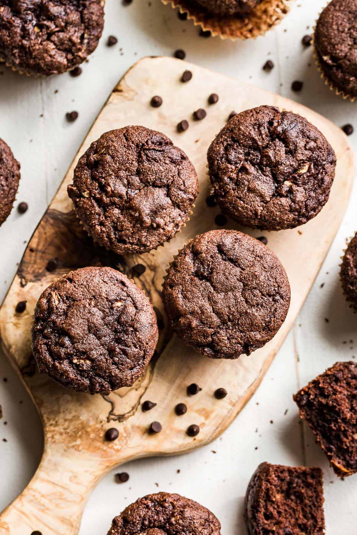 Paleo Chocolate Zucchini Muffins on a wood cutting board with chocolate chips scattered around them and one muffin broken in half.