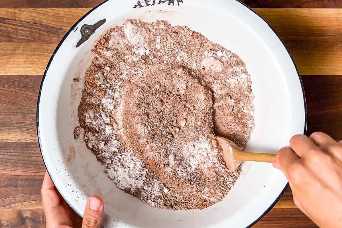 Mixing together the dry ingredients for the healthy chocolate zucchini muffins in a large white bowl on a wood cutting board.