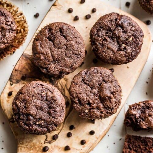 Healthy Chocolate Zucchini Muffins on a wood cutting board with chocolate chips scattered around them.